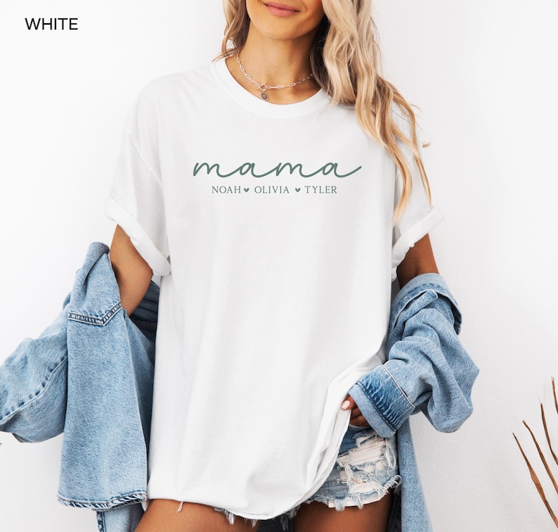 May include: White t-shirt with the word "mama" in a script font, followed by the names Noah, Olivia, and Tyler. The shirt has rolled-up sleeves and is paired with a denim jacket and shorts.