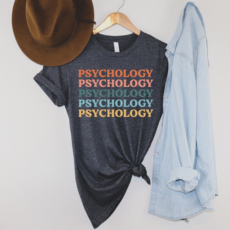 May include: A dark grey t-shirt with the word "Psychology" repeated five times in a rainbow color pattern. The shirt is on a hanger and has a knotted bottom.