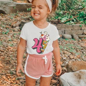 Pode incluir: Uma menina pequena vestindo uma camiseta branca com um gr&aacute;fico de cone de sorvete rosa e branco que diz "4". Ela tamb&eacute;m est&aacute; vestindo shorts rosa com detalhes brancos e um la&ccedil;o rosa no cabelo.