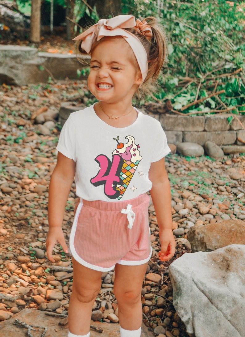 Pode incluir: Uma menina pequena vestindo uma camiseta branca com um gr&aacute;fico de cone de sorvete rosa e branco que diz "4". Ela tamb&eacute;m est&aacute; vestindo shorts rosa com detalhes brancos e um la&ccedil;o rosa no cabelo.