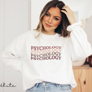 May include: White sweatshirt with the word "Psychology" repeated four times in a stacked, retro-style font. The text is in shades of pink and brown.