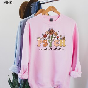 May include: Pink sweatshirt with a floral design and the text "PSYCH nurse" in a retro font.