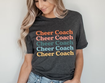 May include: A dark grey t-shirt with the text "Cheer Coach" repeated five times in a rainbow of colors. The text is stacked vertically.