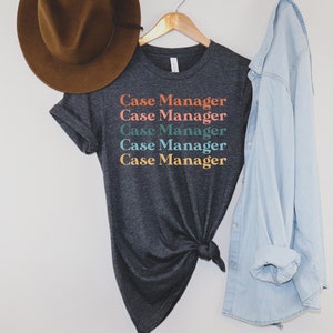K&ouml;nnte beinhalten: Ein dunkelgraues T-Shirt mit dem Text "Case Manager", der f&uuml;nfmal in verschiedenen Farben wiederholt wird. Der Text ist vertikal gestapelt, wobei jede Zeile in einer anderen Farbe ist: Orange, Rosa, T&uuml;rkis, Blau und Gelb.