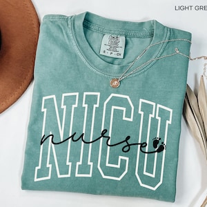 May include: Light green t-shirt with the text "NICU nurse" in white and black lettering. The text "NICU" is in a large, bold font with a white outline. The word "nurse" is written in cursive with a black outline. There are two small black footprints at the end of the word "nurse".