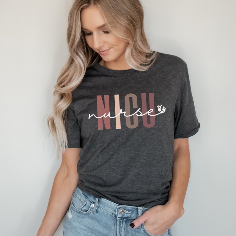 Op de afbeelding: Een donkergrijs T-shirt met de tekst "NICU nurse" in witte letters. De letters "NICU" zijn in een kleurrijk bloklettertype met roze, perzikkleurige en bruine kleuren. Naast het woord "nurse" staan twee kleine witte voetafdrukken.