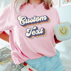May include: A pink t-shirt with a retro-style graphic that says "Custom Text" in rainbow colors.