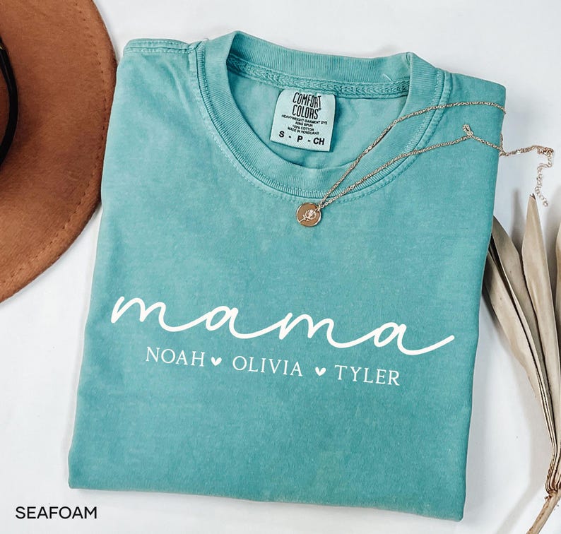 May include: Seafoam green t-shirt with the word "mama" in white script. Below the word "mama" are the names "Noah", "Olivia", and "Tyler" with heart symbols. A gold necklace with a pendant rests on the shirt.