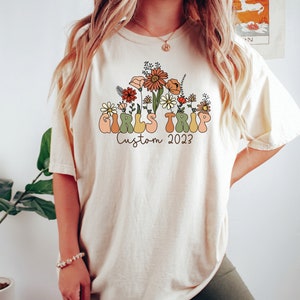 May include: A white t-shirt with a graphic design of flowers and the text "Girls Trip Custom 2023".