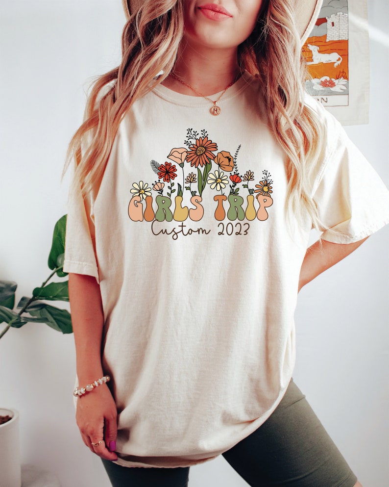 May include: A white t-shirt with a graphic design of flowers and the text "Girls Trip Custom 2023".