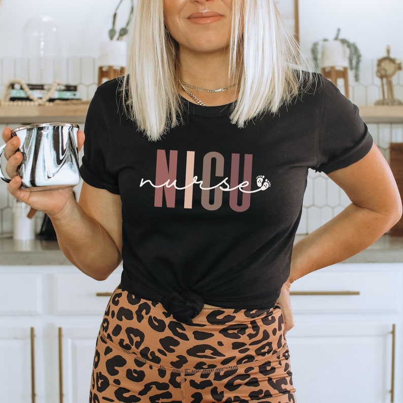 Op de afbeelding: Zwart T-shirt met de tekst "NICU nurse" in roze, bruine en witte letters met een kleine voetafdruk. Het shirt is in de taille geknoopt en de persoon die het draagt ​​draagt ​​ook een luipaardprintbroek.