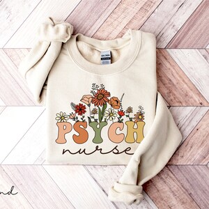 May include: A cream-colored sweatshirt with a floral design and the text "PSYCH nurse" in colorful, retro-style lettering.