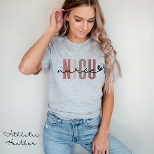 Op de afbeelding: Een grijs T-shirt met de tekst "NICU nurse" in roze, bruine en beige strepen met een kleine voetafdruk. Het shirt is gemaakt van atletisch heather-stof.