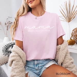 May include: A light pink t-shirt with the word "mama" in a cursive font. Below the word "mama" are the names Noah, Olivia, and Tyler. The shirt is made of a soft material.