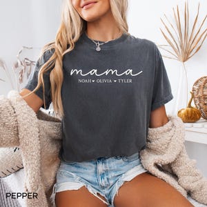 May include: Dark gray t-shirt with the word "mama" in white script. Below the word "mama" are the names "Noah", "Olivia", and "Tyler". The shirt is paired with denim shorts and a cream-colored cardigan.
