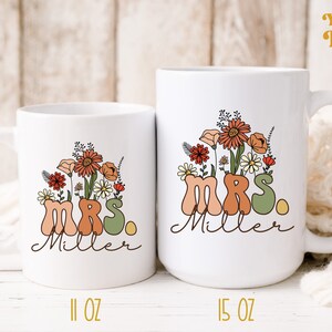 May include: Two white ceramic mugs with a floral design and the text "MRS. Miller" in a retro font. The smaller mug is 11 oz and the larger mug is 15 oz.