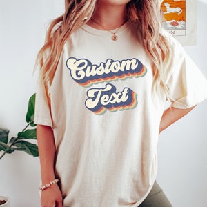 May include: A white t-shirt with a retro-style graphic that says "Custom Text" in a rainbow of colors. The shirt is made of a soft, comfortable material and is perfect for everyday wear.
