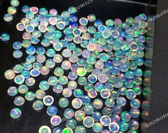 Op de afbeelding: Een close-up afbeelding van een grote hoeveelheid regenboogkleurige opaal cabochons. De cabochons zijn rond en hebben een verscheidenheid aan kleuren, waaronder wit, blauw, groen en geel. De cabochons zijn willekeurig gerangschikt op een zwart oppervlak.