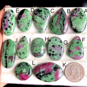 May include: A selection of polished ruby in zoisite gemstones in diverse shapes, such as oval and teardrop. The stones exhibit a blend of green, black, and pink colours. A coin is included for size comparison.
