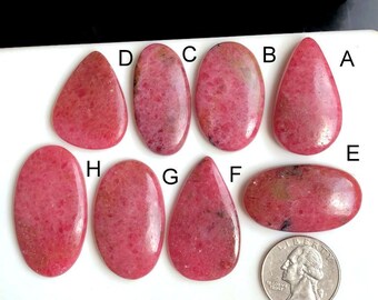 May include: A collection of eight polished, pink rhodochrosite gemstones in various shapes, including oval, teardrop, and irregular forms. The stones display a mottled texture and range in size. A quarter coin is included for size reference.