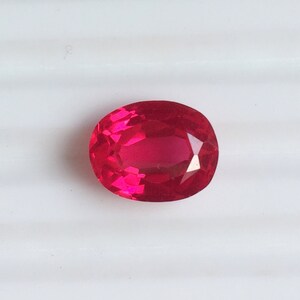 Pode incluir: Uma gema vermelha vibrante, facetada e de formato oval. A gema tem um alto grau de clareza e brilho, refletindo a luz em suas muitas facetas. A gema está sobre um fundo branco.