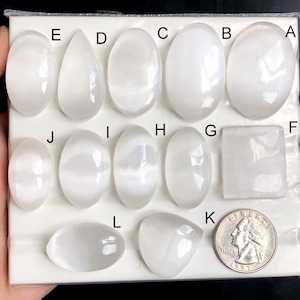 Puede incluir: Una colección de piedras de cristal de selenita blancas en varias formas, incluyendo ovaladas, en forma de lágrima y rectangulares. Se incluye una moneda para la comparación de tamaños. Las piedras están dispuestas sobre una superficie blanca sobre un fondo negro.