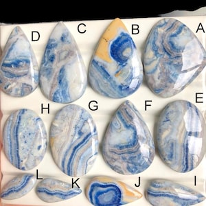 May include: Assortment of polished gemstone cabochons in various shapes, including teardrop and oval. The stones feature a mix of blue, white, and tan colors with unique swirling patterns. The cabochons are displayed on a white surface.