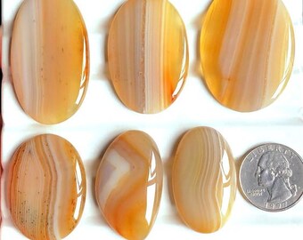 Pode incluir: Seis pedras de ágata ovais polidas em tons de laranja, creme e castanho, com uma moeda para comparação de tamanho. As pedras têm um acabamento brilhante e padrões de bandas naturais. As pedras são exibidas em uma superfície branca.