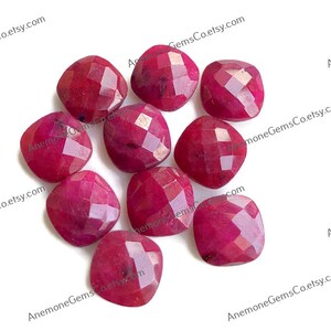 May include: A collection of ten faceted, cushion-cut gemstones in a deep, rich red hue. The gems are arranged on a white surface, showcasing their polished surfaces and intricate cuts. The gemstones are likely rubies.