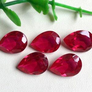 Puede incluir: Cinco gemas facetadas, en forma de lágrima, de color rojo intenso. Las gemas están dispuestas sobre una superficie blanca, con una ramita de hojas verdes en la esquina superior izquierda. Las gemas están talladas para reflejar la luz.