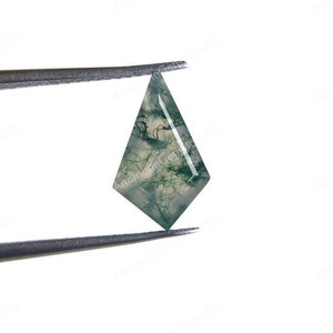May include: A green and white agate gemstone cabochon with a pointed, teardrop shape. The stone is held by tweezers against a white background.
