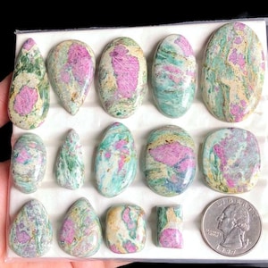 May include: A collection of polished gemstones in various shapes, including oval, teardrop, and rectangular. The stones feature a mix of colors, including pink, green, and cream. A US quarter is included for size reference.