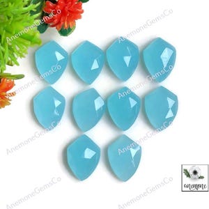 May include: Ten light blue gemstone cabochons, each with a faceted shield shape, are displayed. The gems are arranged on a white surface, with the brand name "Anemone GemsCo" printed around the edges. A small logo is also visible.