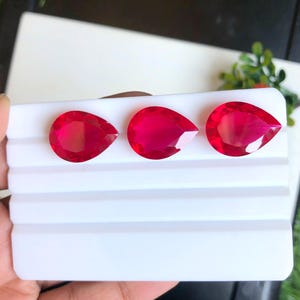 Peut inclure: Trois pierres précieuses rouges en forme de poire sont présentées sur une surface blanche striée. Les gemmes sont facettées, captant la lumière et mettant en valeur leur couleur intense. L'arrangement suggère une présentation de bijoux ou de pierres précieuses.
