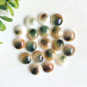 Könnte beinhalten: Eine Sammlung runder, polierter Cabochons in Weiß-, Grün- und Brauntönen. Die Cabochons haben eine glänzende Oberfläche und unterschiedliche Muster, wobei einige einen Farbverlauf aufweisen. Die Anordnung ist verstreut und zeigt die natürlichen Variationen.