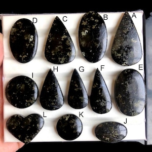 May include: A collection of polished gemstones in various shapes, including oval, teardrop, and heart. The stones are primarily dark green with flecks of gold and are arranged on a white surface. The gemstones are labeled with letters.