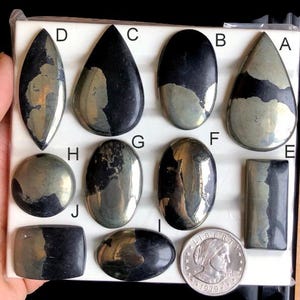 Pode incluir: Uma coleção de pedras preciosas de pirita polidas em várias formas, incluindo gotas, ovais e retângulos. As pedras exibem um brilho metálico com uma mistura de cores cinza escuro e dourado. Uma moeda de 25 centavos de dólar americano é incluída para referência de tamanho.