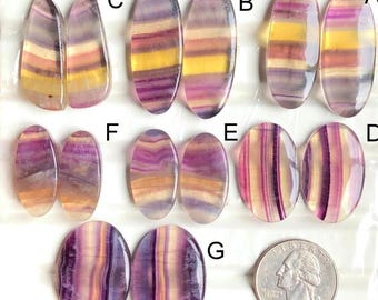 Puede incluir: Una colección de piedras preciosas de fluorita pulidas en varias formas y tamaños, que muestran una mezcla de bandas moradas, amarillas y claras. Las piedras se exhiben sobre una superficie blanca, con una moneda de veinticinco centavos estadounidense para comparar el tamaño. Las piedras son ovaladas y en forma de lágrima.