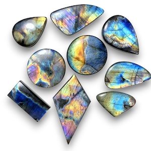 Puede incluir: Una colección de piedras preciosas de labradorita pulidas en varias formas, incluyendo redondas, en forma de lágrima y rectangulares. Las piedras muestran destellos iridiscentes de azul, dorado y púrpura, sobre un fondo oscuro.