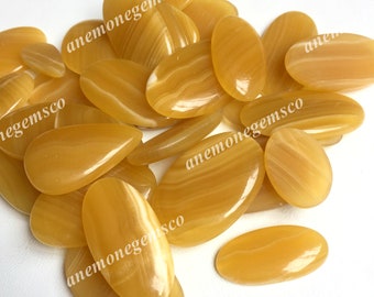May include: A collection of polished, oval-shaped gemstones in shades of yellow and cream. The stones have a smooth, glossy surface with natural banding patterns. The text "anemonegemsco" is printed on some of the stones.
