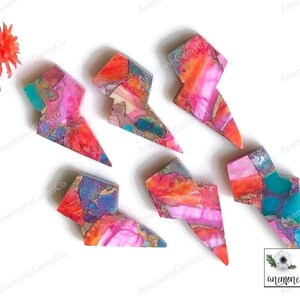 May include: Six colorful gemstone lightning bolt pendants. Each bolt features a unique blend of pink, orange, teal, and gold hues. The pendants are arranged on a white surface, showcasing their vibrant colors and intricate patterns.