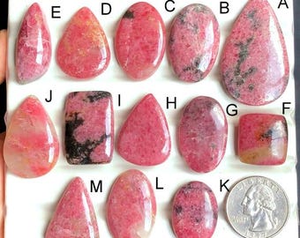May include: Assortment of polished rhodochrosite gemstones in various shapes, including teardrop, oval, and rectangular. The stones display a range of pink and red hues with some black veining. A quarter coin is included for size reference.