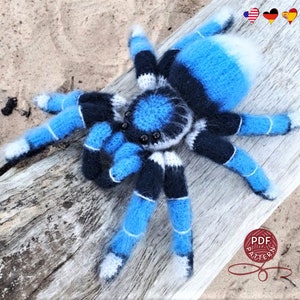 Könnte beinhalten: Eine blaue und schwarze gehäkelte Spinne mit weißen Akzenten. Die Spinne hat acht Beine und einen flauschigen Körper. Die Spinne sitzt auf einem Stück Treibholz.