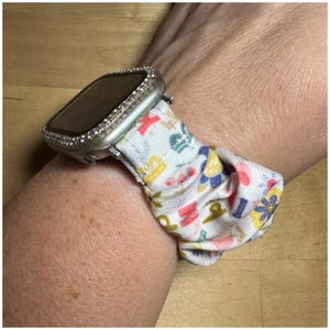 May include: A silver Apple Watch with a clear, sparkly case and a white fabric band with a colorful floral pattern.