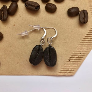 May include: A pair of silver dangle earrings with brown coffee bean charms. The earrings are on a brown paper background with coffee beans scattered around them.