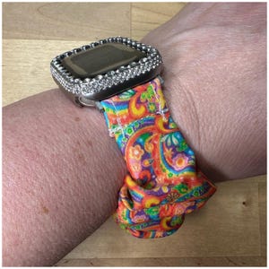 May include: A silver smartwatch with a rhinestone bezel and a colorful paisley patterned band. The band features a vibrant mix of red, orange, yellow, green, and purple hues. The watch is secured on a wrist.
