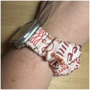 May include: A silver smartwatch with a white and red patterned fabric band. The band is wrapped around the wrist and has the words "Coffee" and "Love" printed on it.