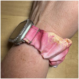 May include: A silver Apple Watch with a pink and yellow tie-dye fabric band. The watch face is covered in rhinestones.