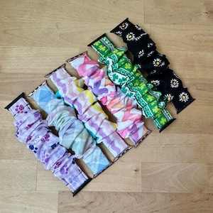 May include: An assortment of fabric watch bands in various patterns and colors. Designs include purple paw prints, blue diamonds, pink and purple waves, watercolor florals, green shamrocks, and black daisies. The bands are displayed on a light wood surface.