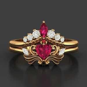 May include: A gold-colored ring set featuring a heart-shaped ruby center stone, surrounded by smaller white gemstones and a marquise-cut ruby. The ring has an ornate, crown-like design.